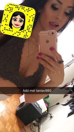 Brunette woman in a black lace top takes a mirror selfie with a rose gold phone, showing her upper body and painted