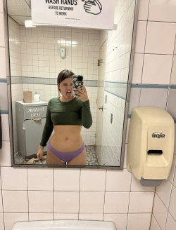 A woman in a green crop top and purple thong takes a selfie in a bathroom.