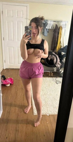 A curvy woman in a black crop top and pink shorts takes a mirror selfie, barefoot in a bedroom with clothes visible in