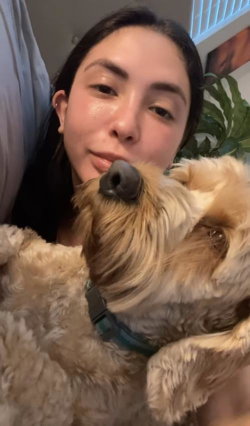 Brunette woman with a fluffy dog, lying close together indoors, smiling softly at the camera.