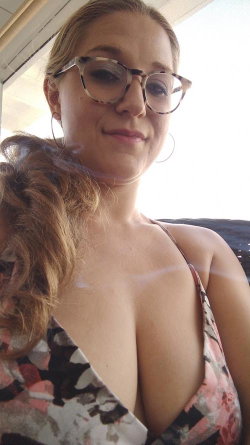 Blonde woman with glasses wearing a floral top, taking a selfie outdoors with sunlight behind her.