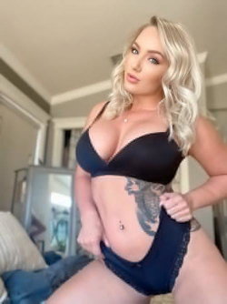 Blonde woman with wavy hair in black lingerie poses indoors, showing a large tattoo on her side and midriff.