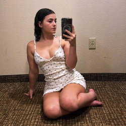 A curvy woman with dark hair sits on a carpeted floor, wearing a white polka-dot dress, taking a mirror selfie with her