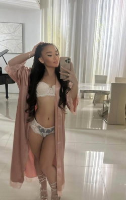 A woman with long dark hair takes a mirror selfie in a white lace bra and patterned underwear, wearing a pink robe and