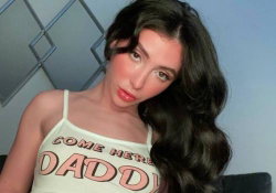 Brunette woman with long wavy hair wearing a white tank top with text, posing indoors near a geometric wall design.
