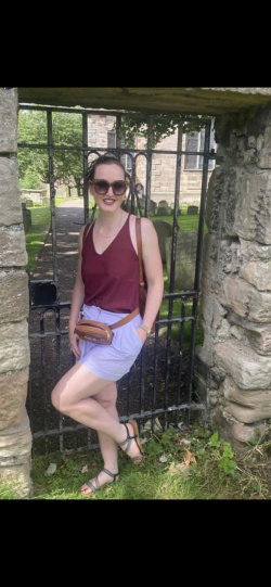 A woman with dark hair and sunglasses poses in a maroon tank top and light blue shorts, standing by a stone gate in a