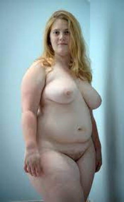 A blonde woman in a thong stands topless in a plain room.