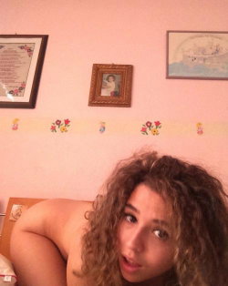 Curly-haired woman with bare shoulders lying on a bed in a room with pink walls and framed pictures.