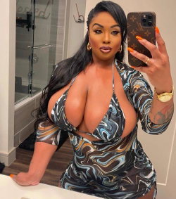 A curvy woman with long black hair takes a mirror selfie in a patterned dress, showcasing her figure in a bathroom
