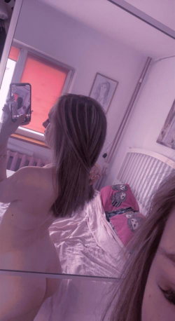 A woman with shoulder-length brown hair takes a mirror selfie in a bedroom, topless, wearing light pink pajama bottoms.
