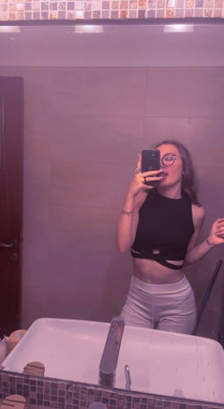Woman in black crop top and white pants taking a selfie in a bathroom.
