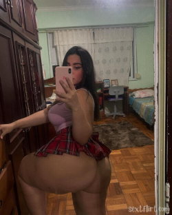 A curvy woman with long dark hair takes a mirror selfie in a pink top and red plaid skirt, standing in a bedroom with