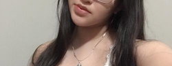 Close-up selfie of a woman with long dark hair, wearing glasses and a delicate necklace, in a neutral indoor setting.