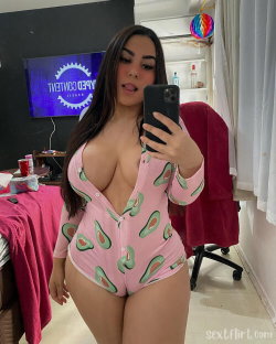 Brunette woman in a pink avocado-print onesie takes a mirror selfie in a bedroom with a TV and desk visible.