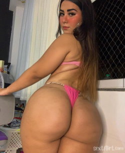 Curvy woman with long dark hair wearing a pink thong and matching top, posing indoors near a window with mesh screen.
