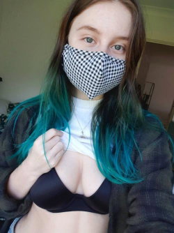 Woman with long brown hair dyed teal at ends, wearing black-and-white checkered face mask, black bra, white crop top,