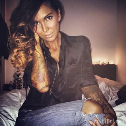 A woman with long wavy hair and visible tattoos on her arms sits on a bed, wearing a dark satin shirt and ripped jeans.