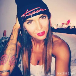 Brunette woman with tattoos on her arm wearing a beanie and white top, posing in bed with flowers in the background.