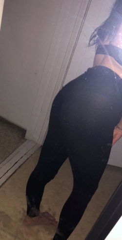 A woman with dark hair in black leggings and a crop top, seen from behind in a dimly lit room, barefoot on carpet.