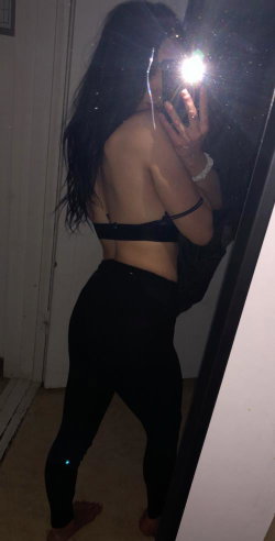 A woman with long dark hair takes a mirror selfie in black lingerie and leggings, showing her back and side profile.