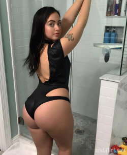 Curvy woman with dark hair in a black cutout bodysuit poses in a bathroom, arm raised, tattoo visible on upper arm.