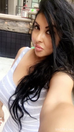 Brunette woman with long dark hair in a striped tank top, taking a selfie outdoors near a stone wall and patio.