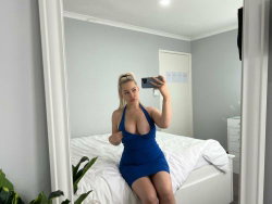 Blonde woman in a blue halter dress takes a mirror selfie while sitting on a bed in a bright bedroom.