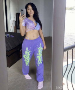 A woman with long dark hair takes a mirror selfie in a purple tie-dye crop top and matching pants with green dragon