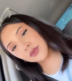 Brunette woman with winged eyeliner and hoop earrings, wearing a white top, taking a selfie in a car.