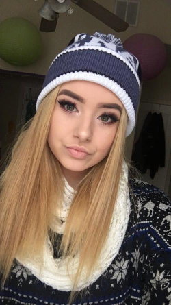 Blonde woman with long hair wearing a blue and white knit beanie, scarf, and sweater in a cozy indoor setting.