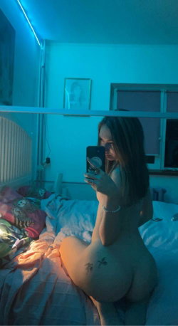 A woman with long dark hair sits nude on a bed in a blue-lit room, taking a mirror selfie with visible tattoos on her