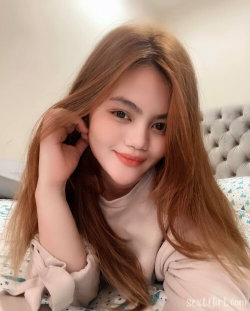 A woman with long light brown hair smiles softly while lying on a bed, wearing a pale pink top with ruffled sleeves.