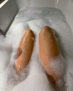 Close-up of legs submerged in bubble bath, skin partially covered in foam, setting appears to be a bathtub.