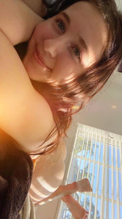 A smiling woman with brown hair and green eyes lies on her side, partially covered by a towel, near a window with