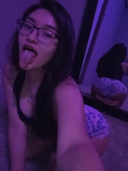 A young woman with long dark hair and glasses sticks her tongue out while wearing a black top and patterned shorts,