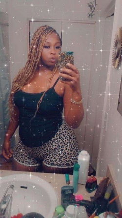 A woman with long braided hair takes a mirror selfie in a bathroom, wearing a black tank top and leopard-print shorts.