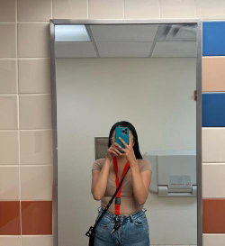 A woman with long dark hair takes a mirror selfie in a beige crop top and blue jeans, holding a phone with a red