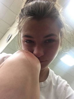 A young woman with light brown hair in a bun, wearing a white t-shirt, takes a close-up selfie indoors with her knee