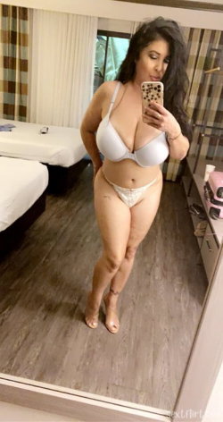 Curvy woman with long dark hair takes mirror selfie in white lingerie, standing in a hotel room with beds and patterned