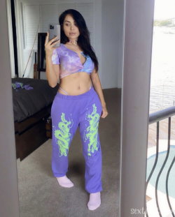 A woman with long dark hair takes a mirror selfie in a tie-dye crop top and purple dragon-print pants, standing indoors