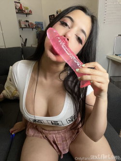 Brunette woman with long dark hair sits on a couch, wearing a white crop top and plaid shorts, holding a pink toy to