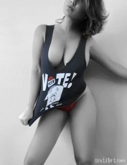 Curvy woman in a low-cut tank top and red underwear poses against a plain wall, lifting her shirt slightly.