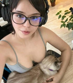 A young woman with short dark hair and glasses wears headphones and a gray tank top while holding a small dog indoors.