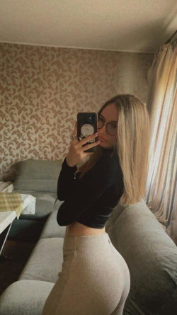 Blonde woman in black crop top and beige high-waisted pants takes a mirror selfie in a living room with patterned