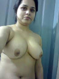 A woman with dark hair and a curvy build stands topless in front of a door, looking directly at the camera.