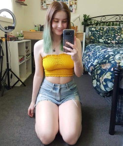 A young woman with pastel green-tipped hair kneels on the floor in a bedroom, wearing a yellow crop top and denim