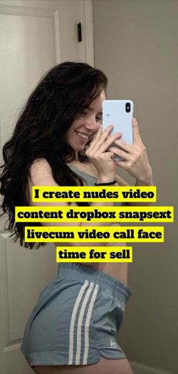 Brunette woman with long curly hair takes a mirror selfie in light blue shorts and a crop top, smiling while holding a