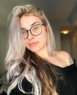 A woman with long, wavy blonde and brown hair, wearing black-rimmed glasses and an off-the-shoulder black sweater,