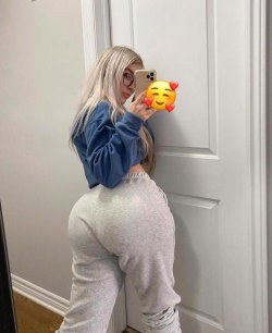 Blonde woman in a blue crop sweatshirt and gray sweatpants takes a mirror selfie, showcasing a curvy figure in a home