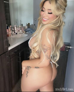 Blonde woman with long wavy hair and visible tattoos poses in a bathroom, wearing a white top and thong, showing curvy